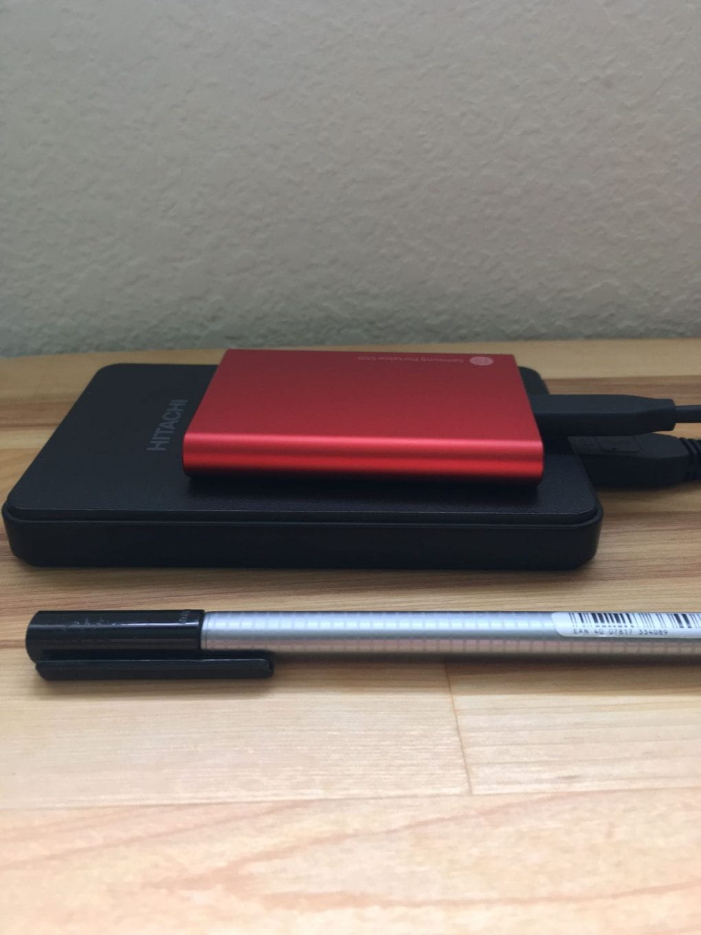 Stacked on top of eachother. On table a pen, Bottom a black spinning disk external HDD, top bright red sold state HDD