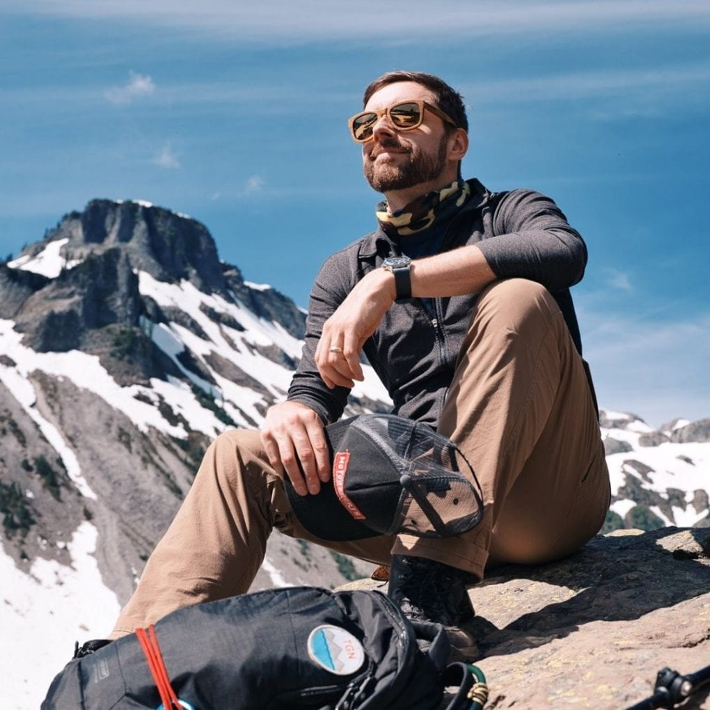 AJ on a mountain with a mountain behind him holding his black hiking had and his camera bag in front of him