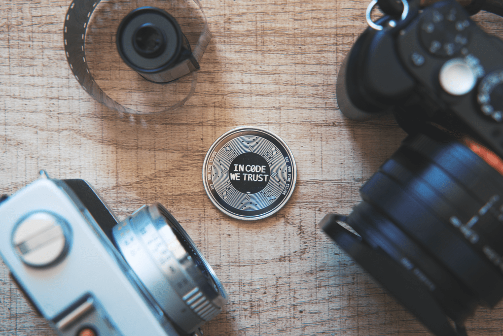 A silver crypto currency coin with the words "In Code We Trust" on a wood table with two cameras and a roll of film