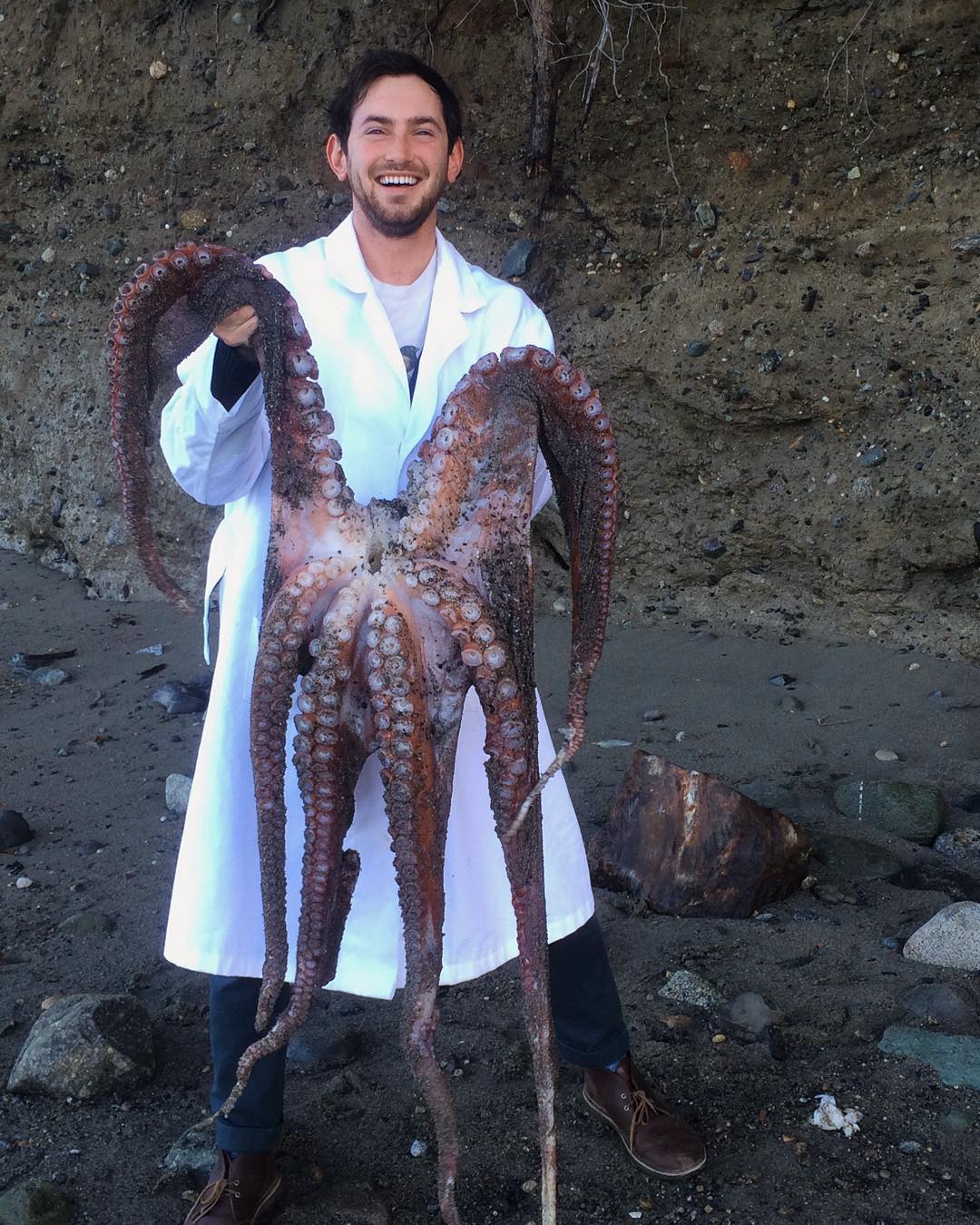 Jake Lawlor smiling, standing on a rocky beach, wearing a white lab coat, and holding a dead giant pacific octopus.