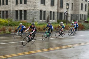 Pack of riders cycle past a building on a grey, wet day