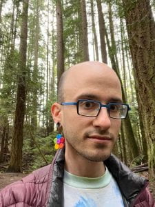A selfie photo of a white man with a rainbow-colored flower earring and blue-rimmed glasses. He is wearing a jacket, and standing with a cloudy forest of trees in the background.