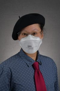 A nonbinary person with lighter brown skin wearing a blue shirt with maroon necktie, a white N95 mask, large rectangular glasses, and a black beret cocked to one side, looking toward the camera.