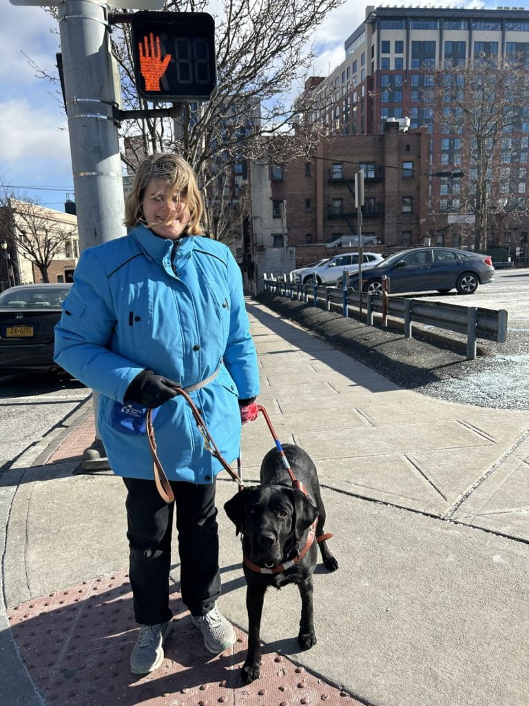 A blonde-haired woman in a sky blue winter coat is standing at a city crosswalk and smiling with eyes closed with a black guide dog at her side.