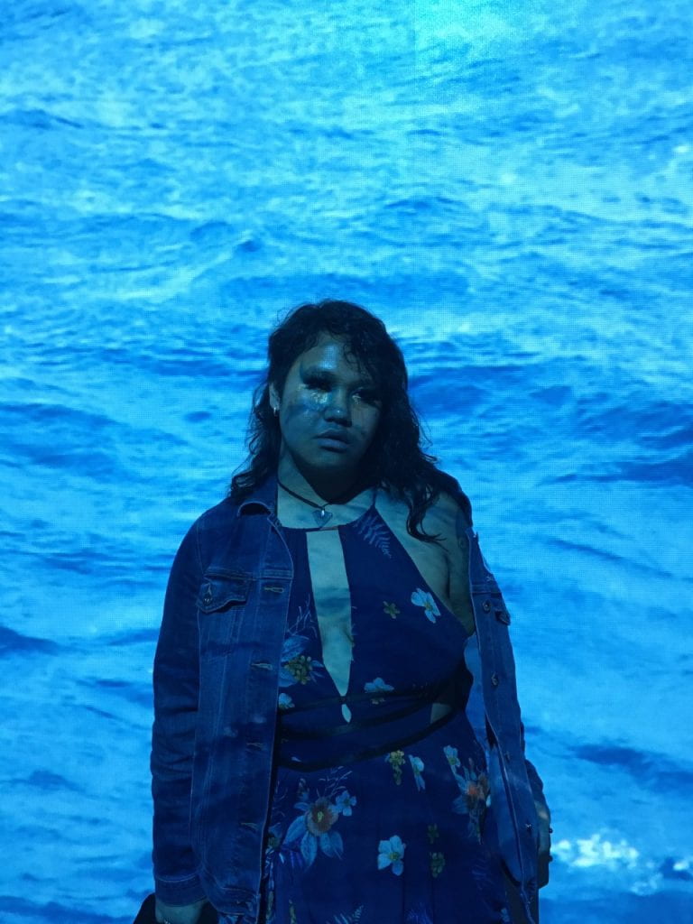 A photo all in tones of blue. A person with dark skin and long, dark hair is wearing a blue floral dress and denim jacket and standing in front of an ocean backdrop that is projecting shades of blue on their face and skin as well.