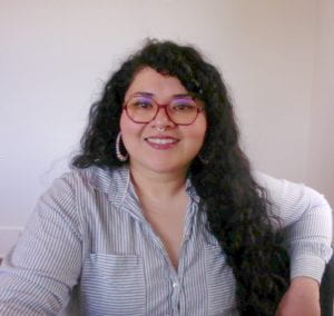 A selfie of a feminine person sitting and smiling at the camera, with magenta-rimmed glasses, pearl earrings, long wavy black hair, and grey and white striped button shirt.