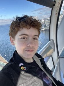 Photo of Jordan Van Haften in the form of a selfie of a light-skinned nonbinary person with freckles and short curly hair. They are in front of a sunny sky and blue water. There are wearing a button denim shirt and have sunglasses resting on top of their head.