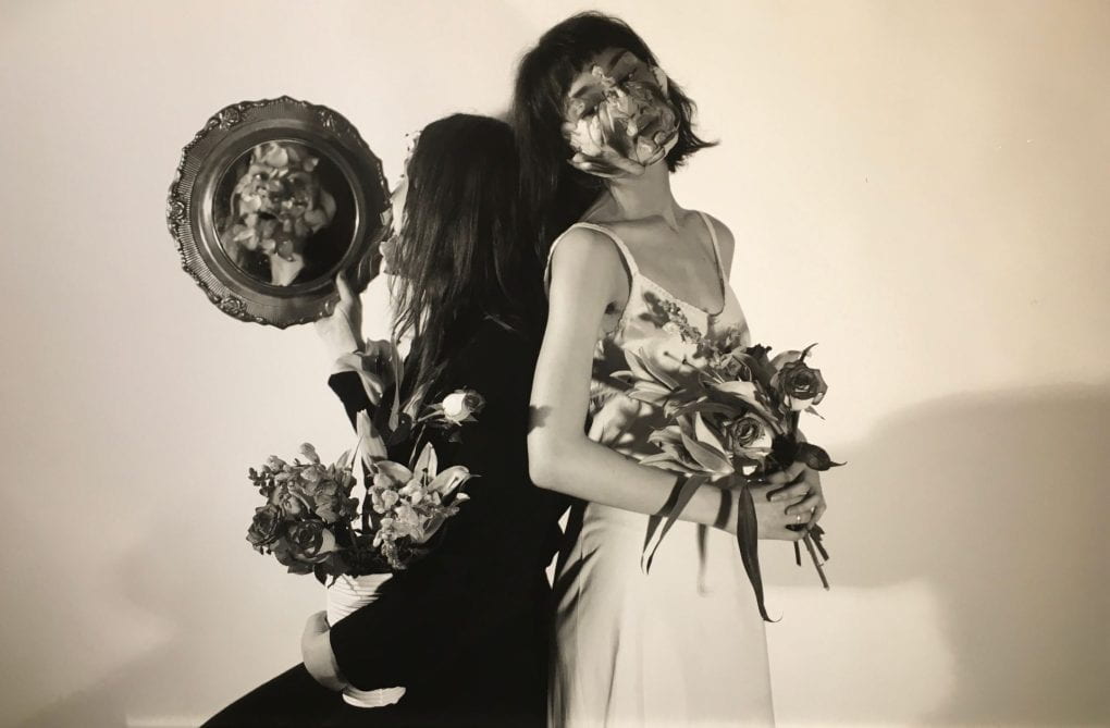 Two women stand back to back with one in a white dress facing the camera showing her face covered in an opaque flower and the other women dressed in black facing away from the camera. They both hold bouquets of flowers, and the women in the black dress whose face would be reflected in a mirror is obscured in the reflection by the flowers at her waist in her hands