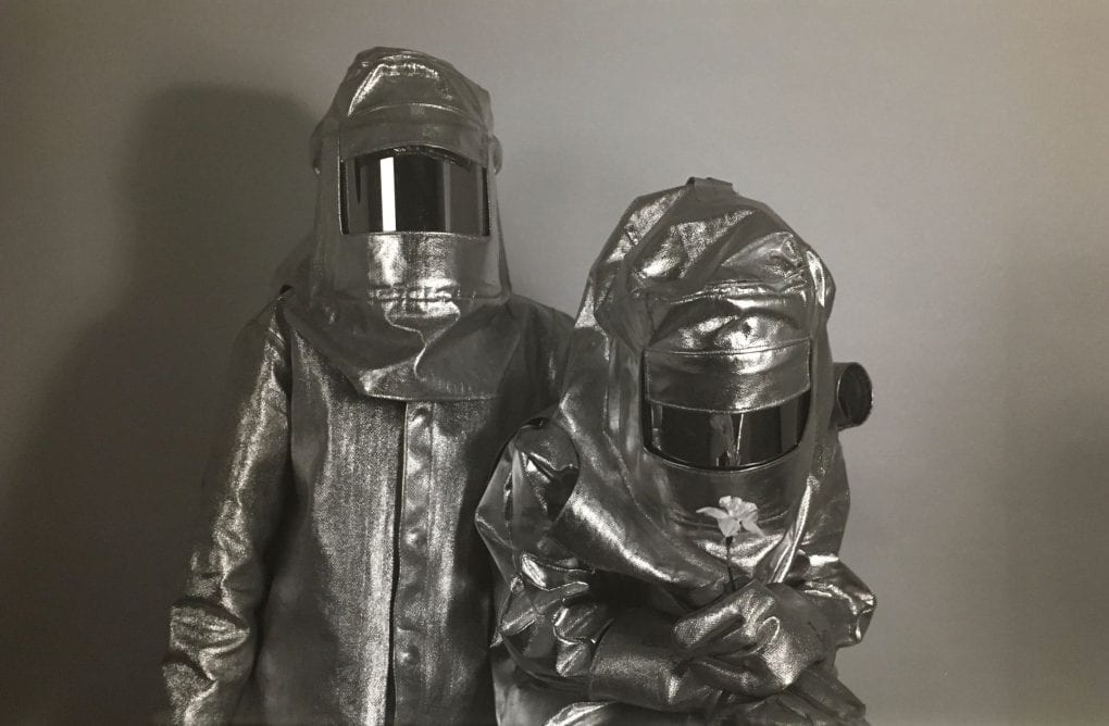 Two figures pose in advanced proximity heat protecting suits, which are full-body covering, are a reflective silver and have rectangular tinted face guards. One figure stands upright and the other is leaning over as if looking at something on the ground