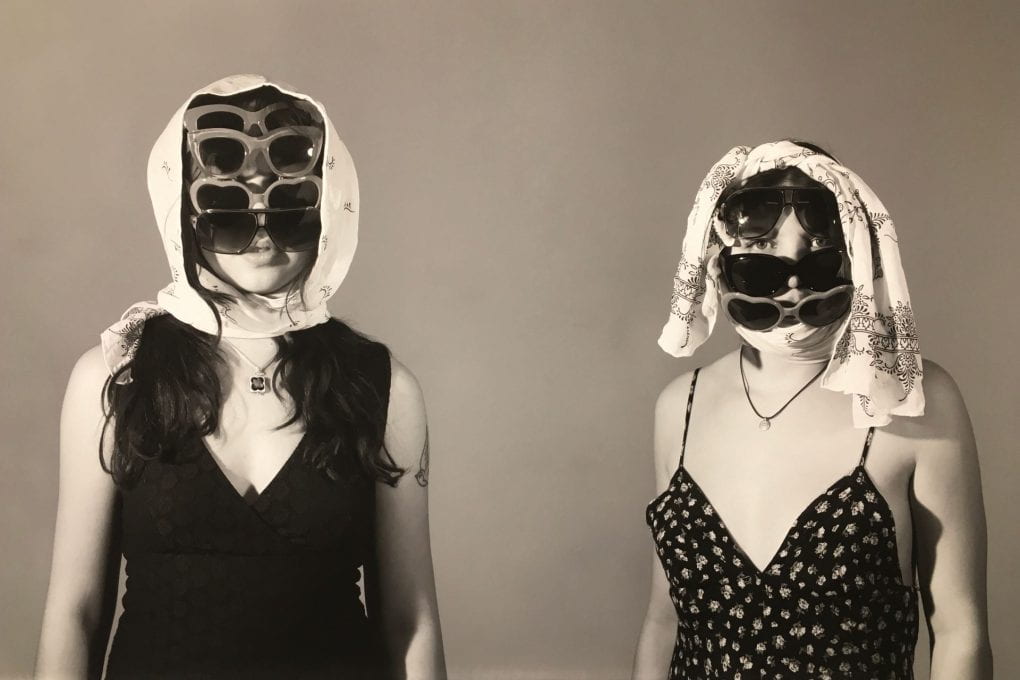Two women look at the camera, both are wearing dark dresses. They have stacked multiple ayers of sunglasses over their face to the point of obscurity and on top of their heads are clothes. The woman on the right beaks through a gap in their shield of sunglasses.