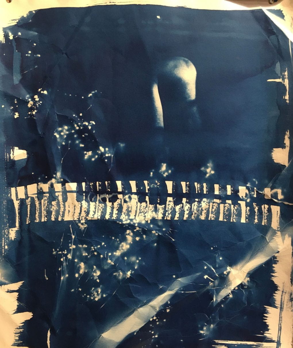 In dark blue cyanotype, an abstract collage piece with a shadowed out figure's shoulder and a streak of piano keys across the page. The paper has been purposely crumbled then re-flattened
