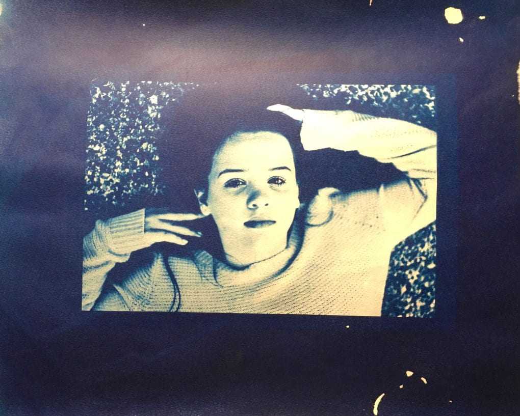 In cyanotype, a person lays on the ground toughing the top of their head and their earlobe, they look at the camera