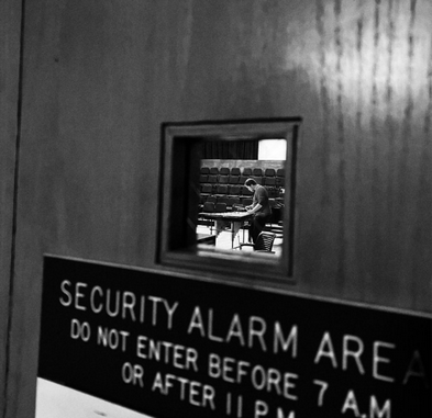 The outside of a door with a sign that reads "security alarm area, do not enter before 7am", through the window of the door in a lecture hall a person organizes papers on a desk