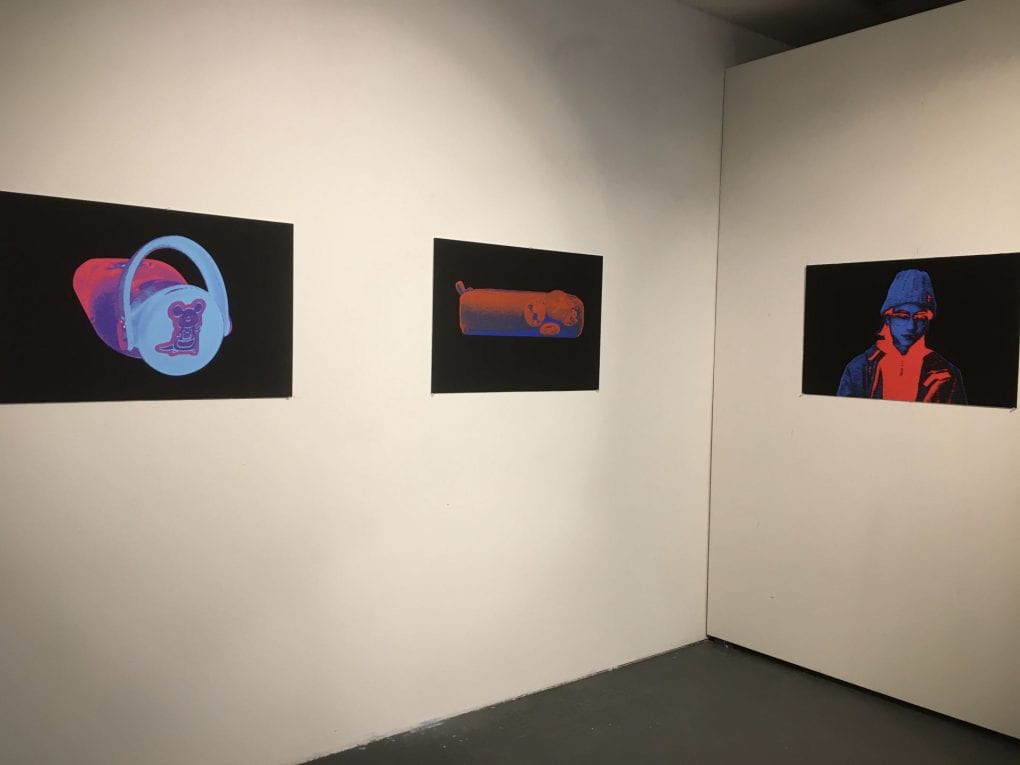 Multiple large black prints set up around a gallery with white walls, the images pop brightly with color, and feature either human portraits or still life's of objects