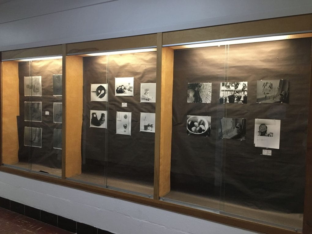 A view of class cases on a wall, each of which contain size medium sized black and white photo prints