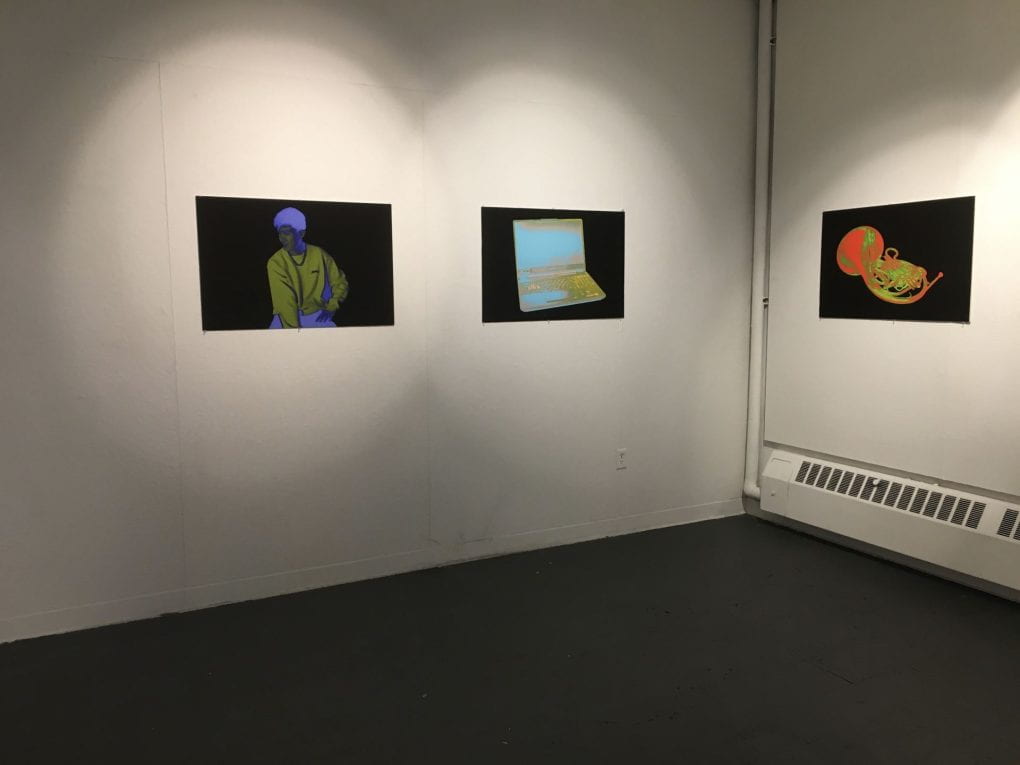 Multiple large black prints set up around a gallery with white walls, the images pop brightly with color, and feature either human portraits or still life's of objects
