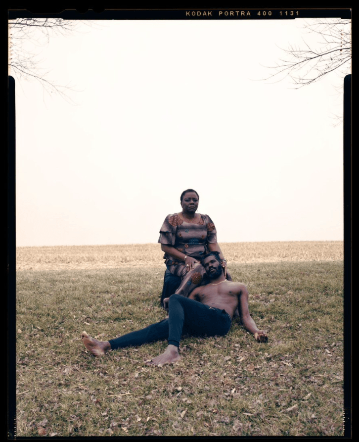 A black woman sits in a harvested, dry field, her adult son rests his head in her lap with his eyes closed, shirtless. Her hand frames his face, she frowns and looks at the audience.