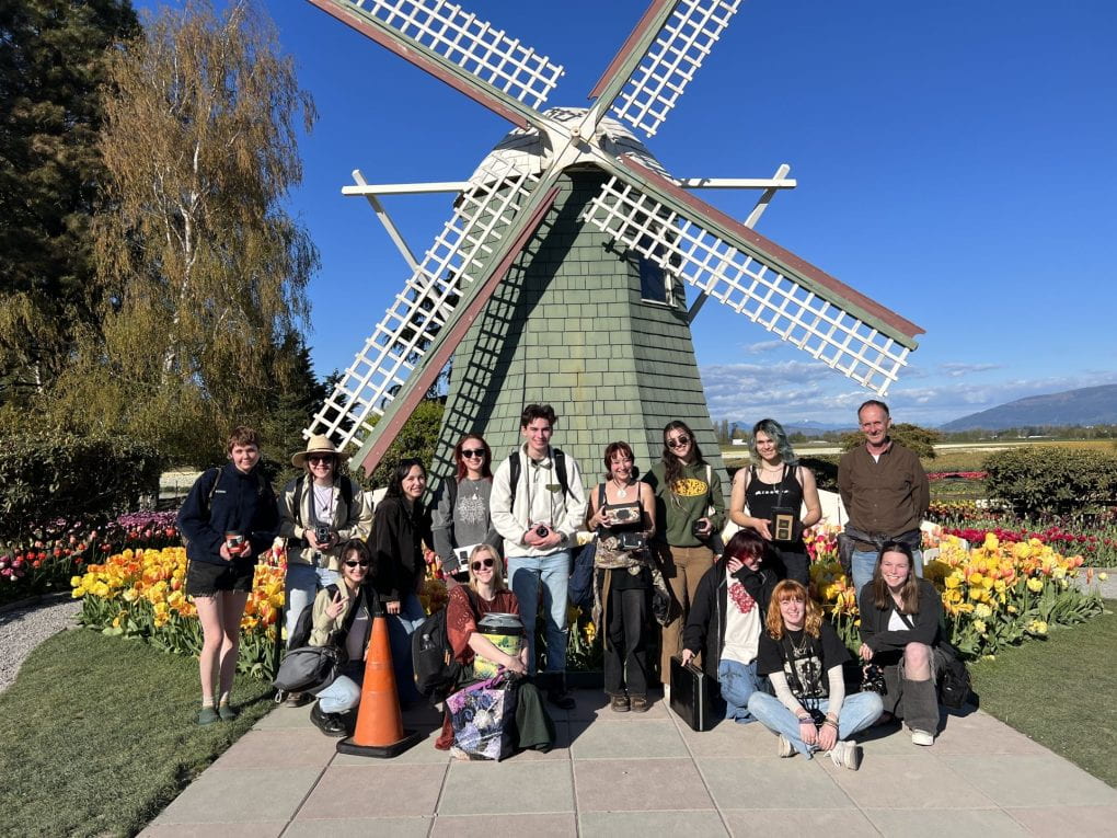 13 students and Nathan Cranston pose with pinhole cameras in front of a green-tiled windmill, bright yellow and red tulips bloom at the base of the windmill. The sun is shining brightly and the sky is bright and clear.