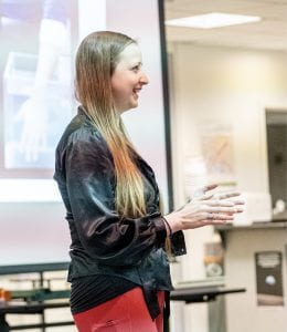 White female Graduate Student, Chrystal Dragonflame, is where black jacket and red pants facing right hands out in the middle of presenting her Three-Minute Thesis.