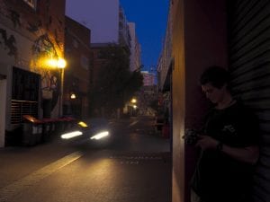 A man with a camera stands to the side of an alley, while a car passes