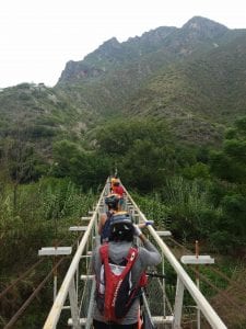 Students cross a narrow suspension bridge into a green mountain