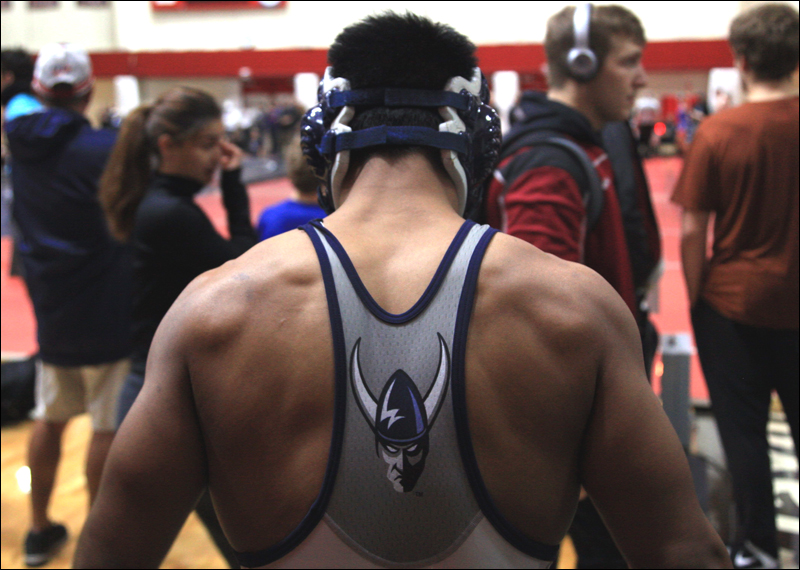 Viking Wrestler stands facing the mat at a competition. Spectators and other competitors stand between him and the mat. 
