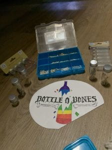 A plastic container and some bottles containing small bones. Bottle O' Bones and a rainbow shattering bottle are drawn on a white paper in fine text.